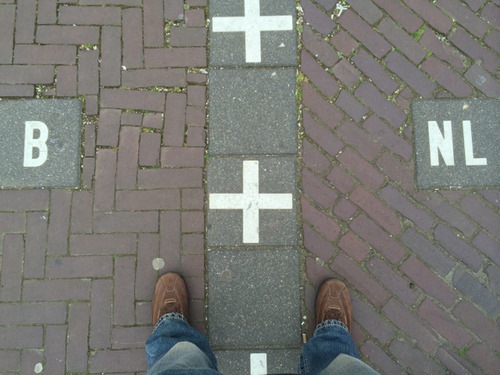Looking down at a pavement where a line of white crosses marks the border, with a B tile on one side and an NL tile on the other