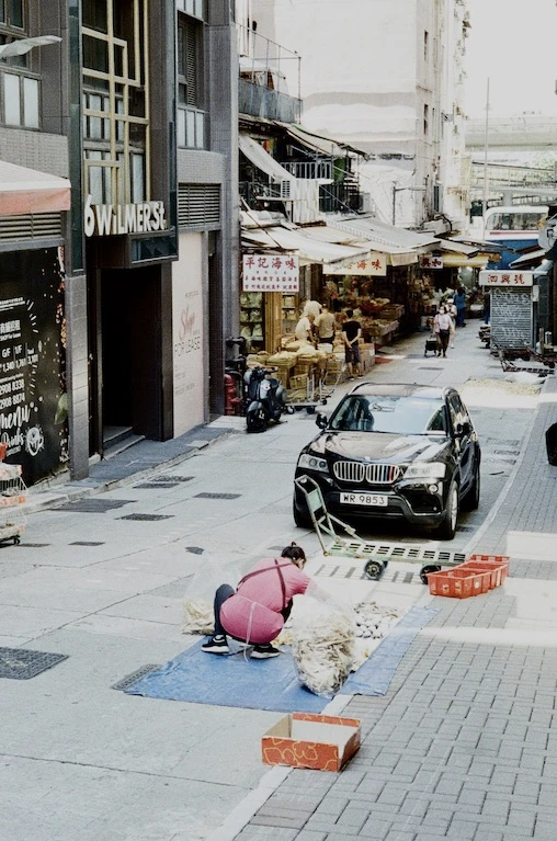 A person sorting goods on the pavement beside a parked luxury car on a Sheung Wan side street