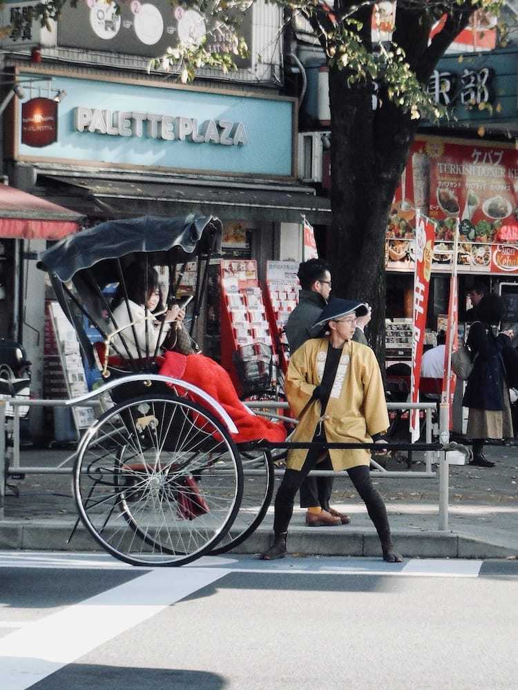 A rickshaw puller in a yellow jacket pulling a red-canopied rickshaw across a crosswalk in Asakusa