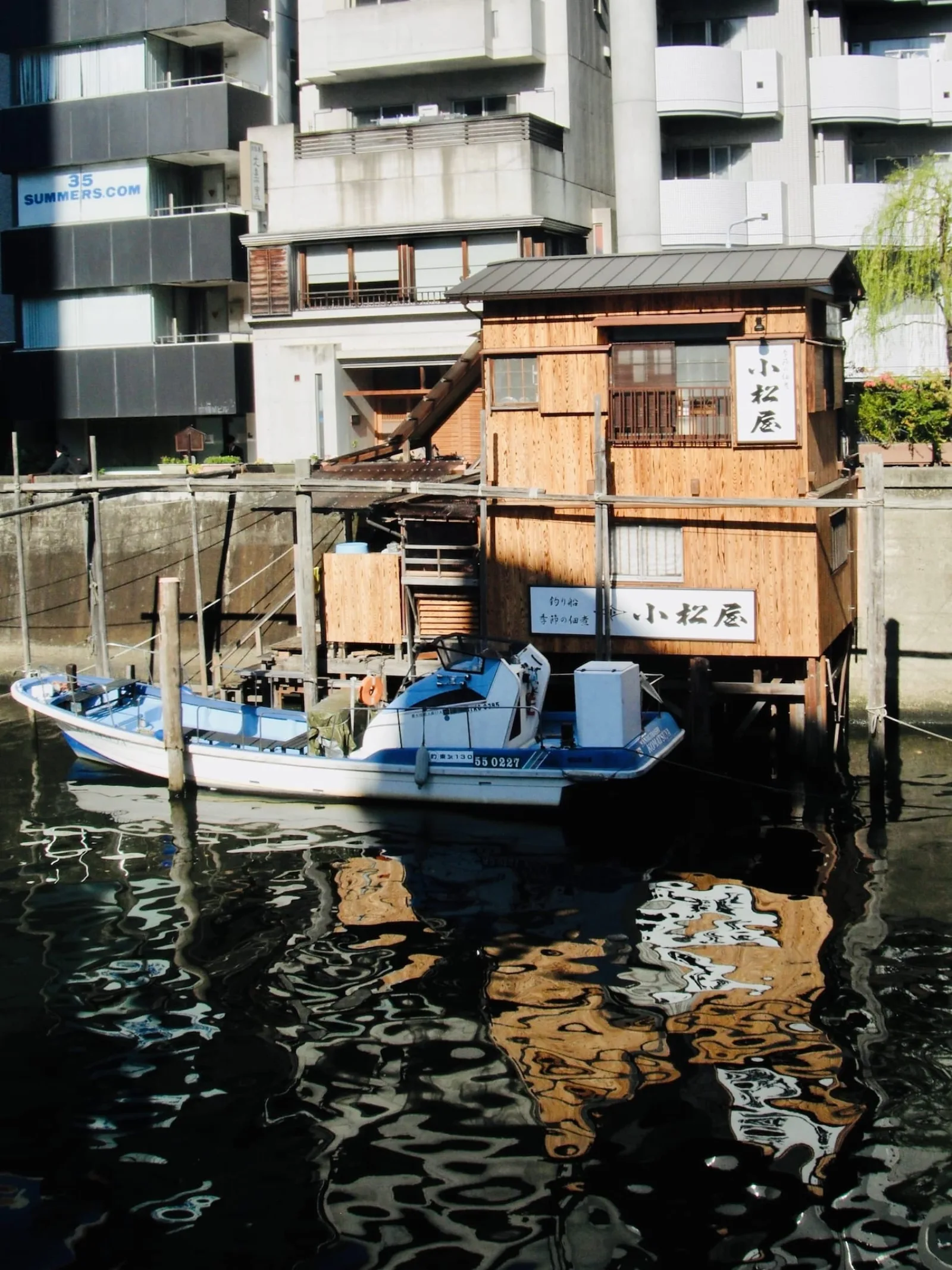 A small fishing boat moored beside a wooden boathouse on a canal, with the buildings reflected in the water