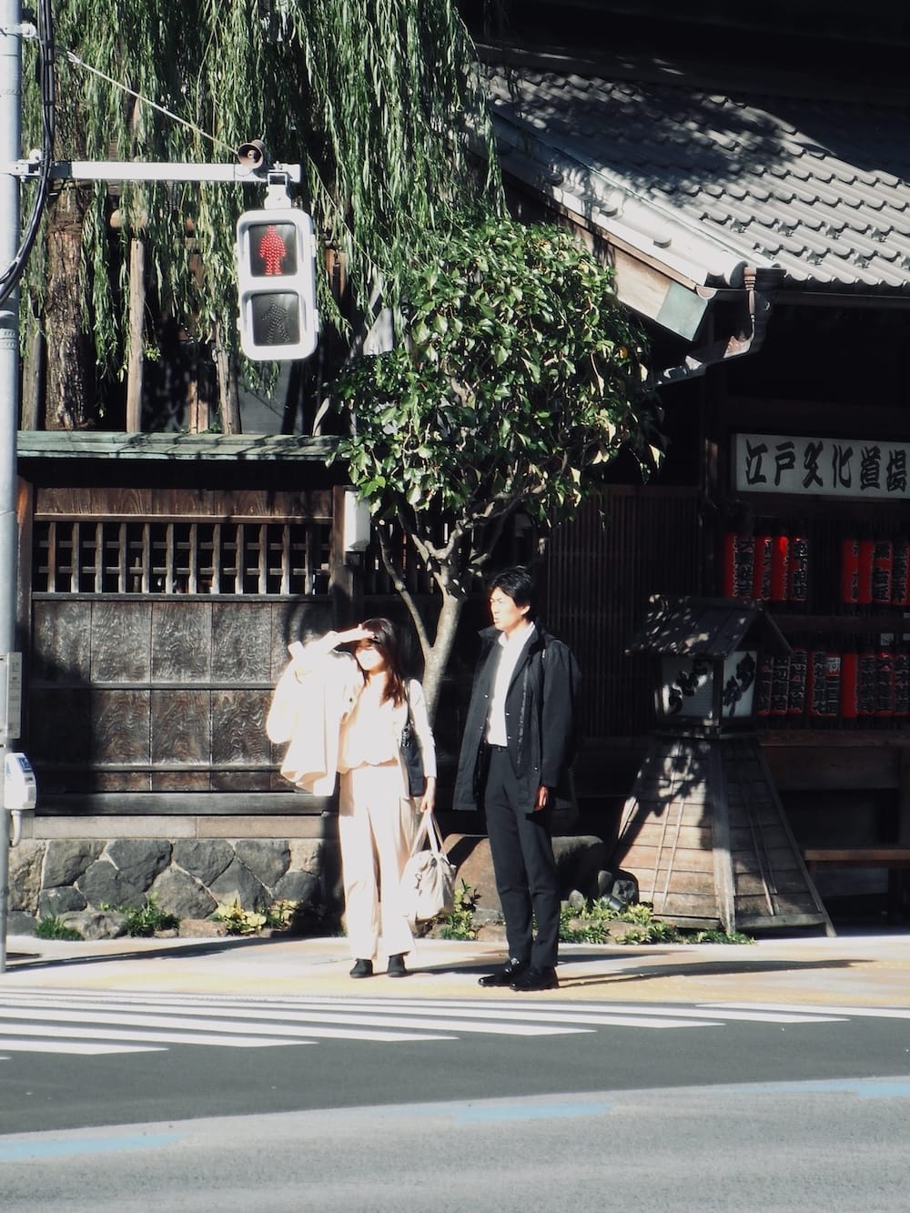Two people waiting at a crosswalk beneath a willow tree outside an old shop