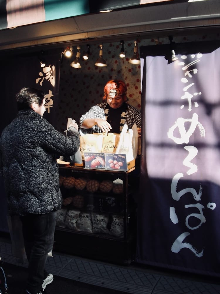 A vendor handing melon pan to a customer at a stall with a purple noren curtain