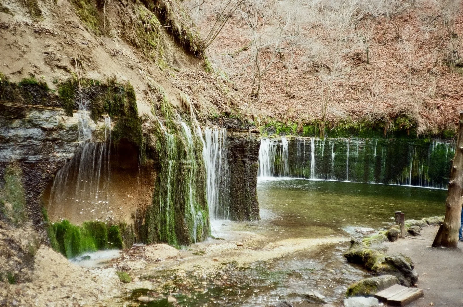 A low, wide waterfall pouring over a mossy rock wall into a pool