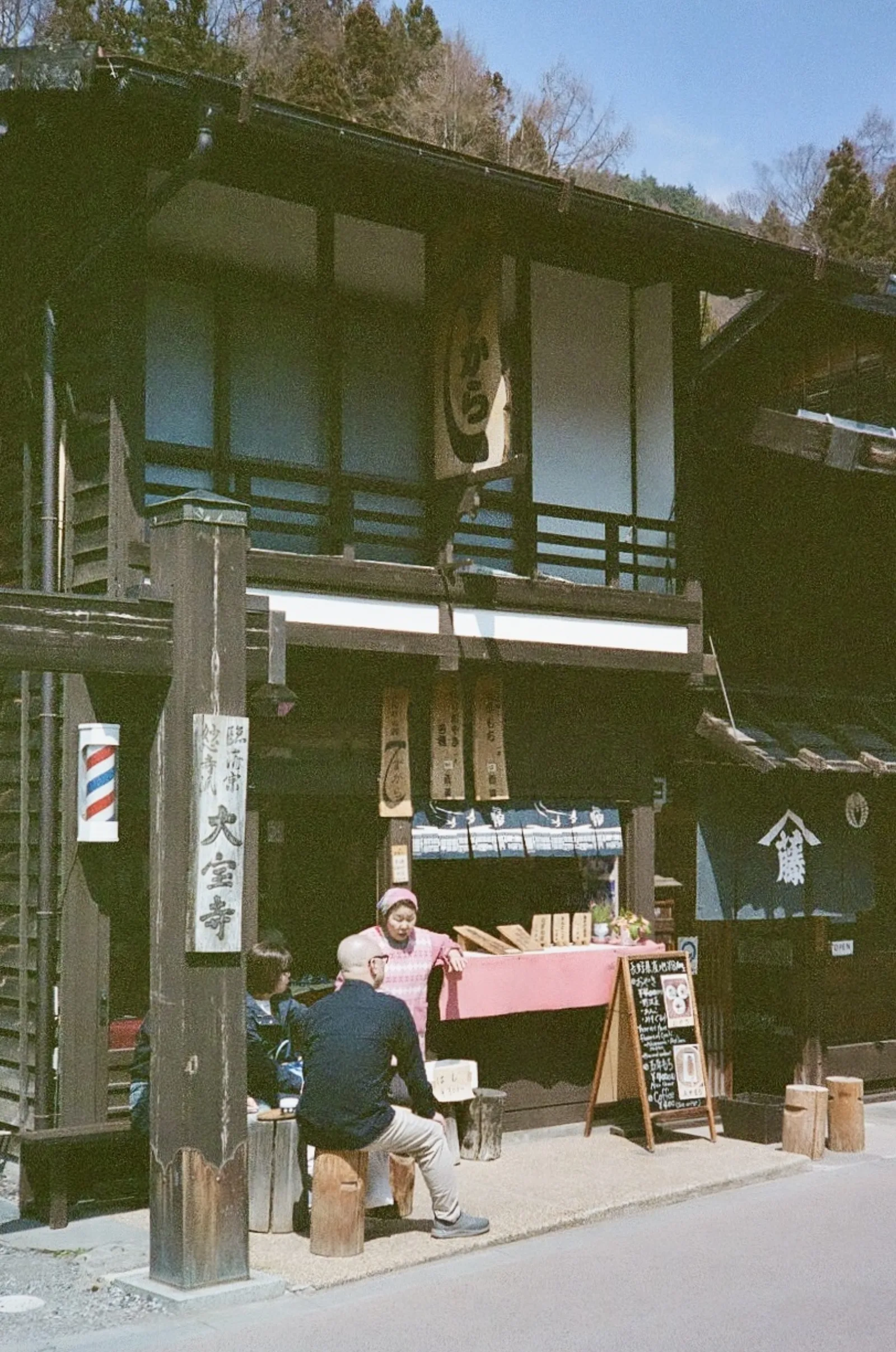 A street vendor seated at a stall outside an old wooden storefront with a barber’s pole