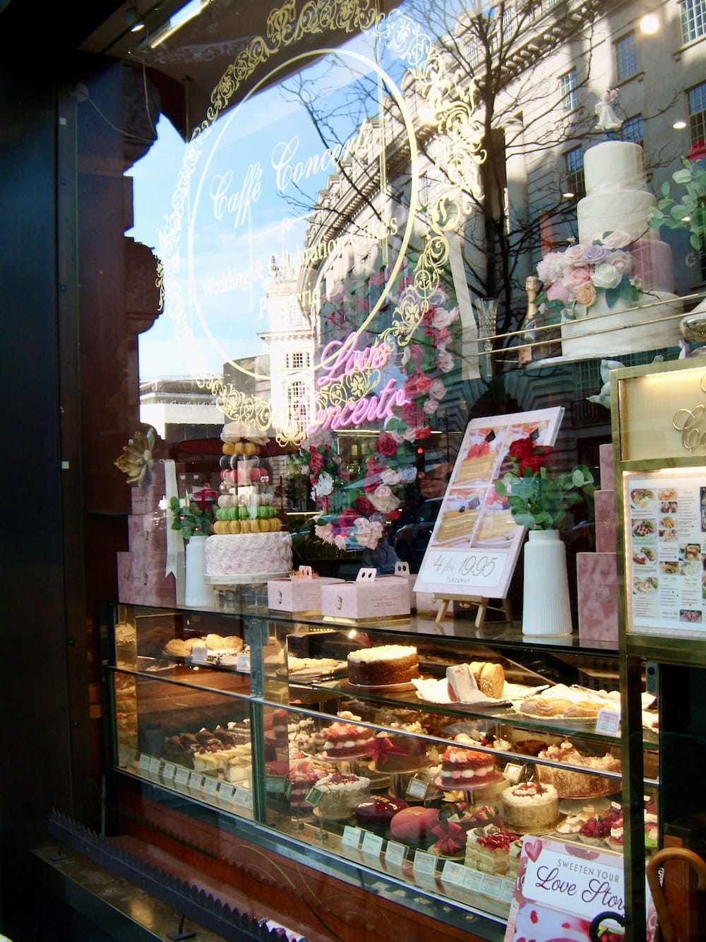 A cake shop window display with tiered cakes, flowers and pastel pastries, and the street reflected in the glass
