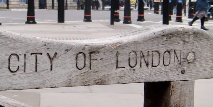 A weathered wooden bench carved with the words City of London, with pedestrians behind