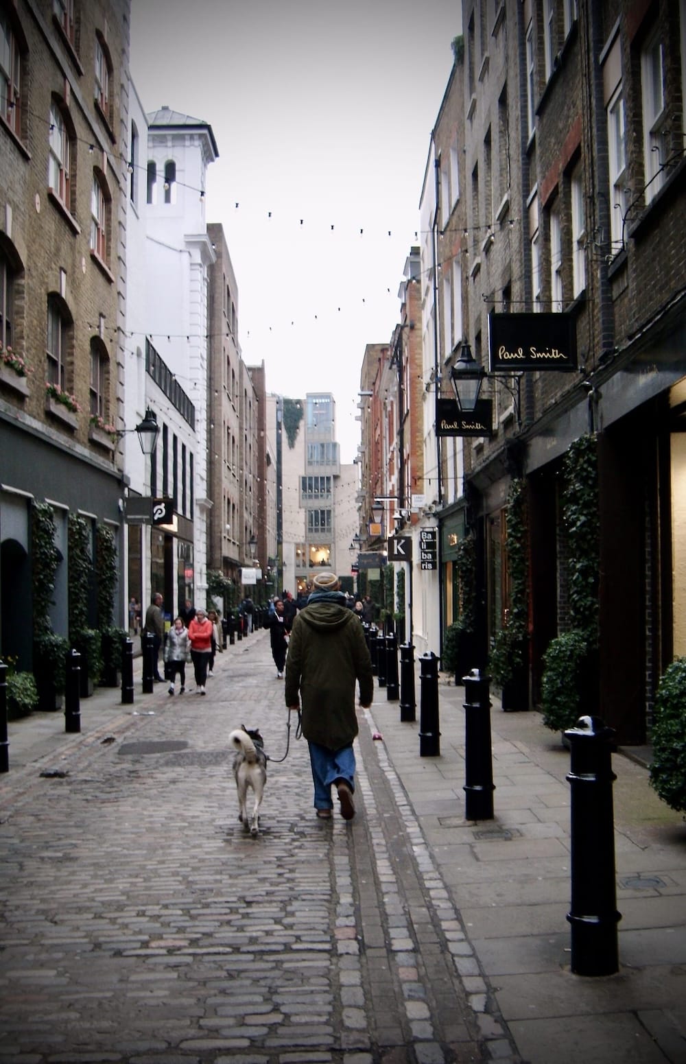 A person walking a dog down a narrow cobbled street lined with shops