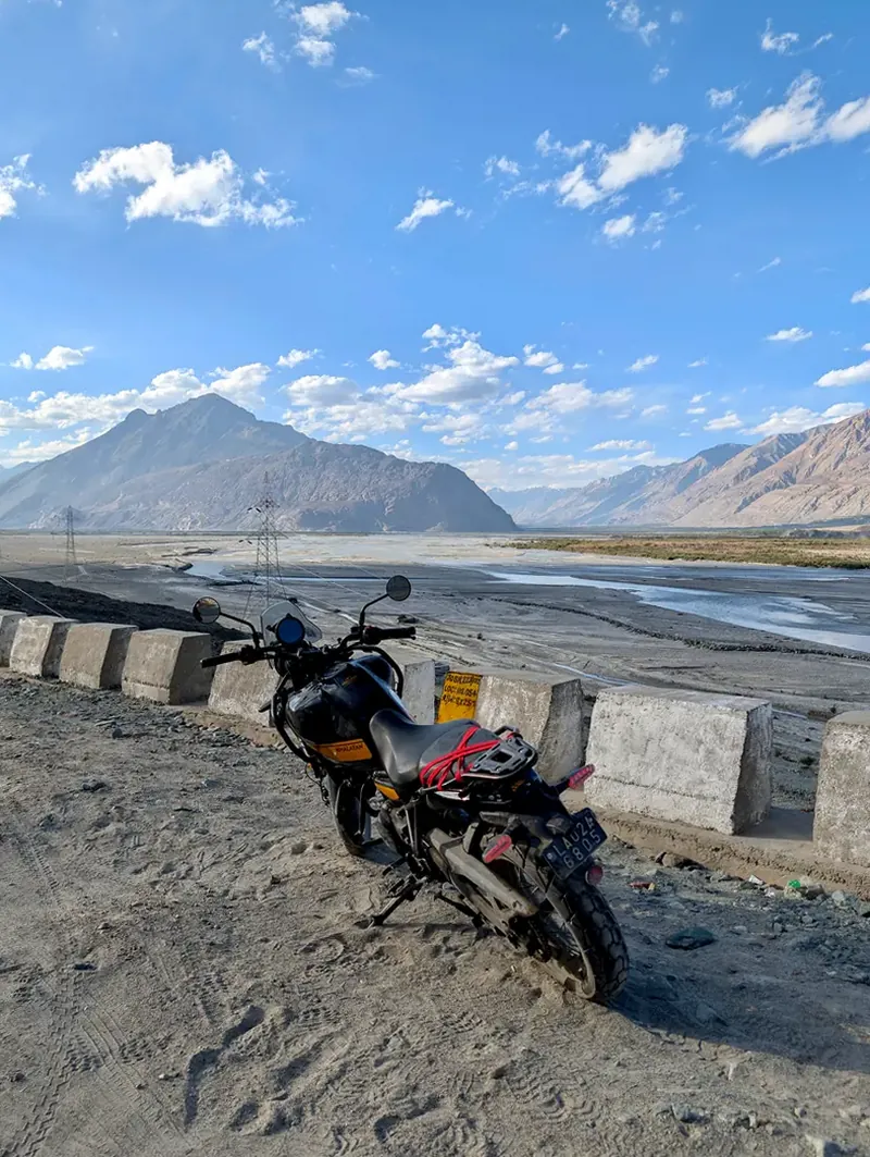 The loaded Royal Enfield Himalayan parked at a low wall above the braided Shyok river in evening light