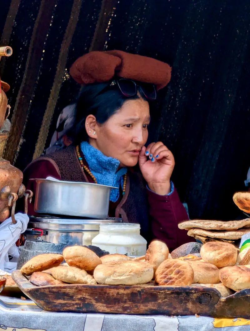 A woman beside a wooden tray piled with round flatbreads and a pot of butter tea