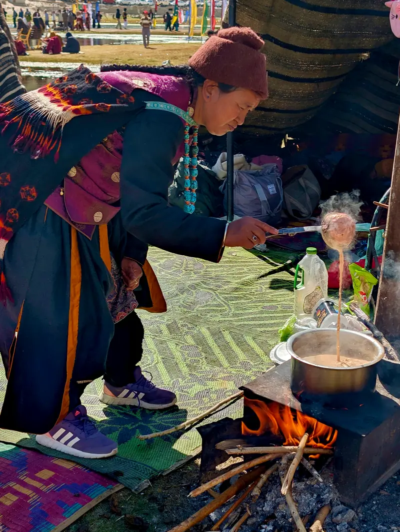 A woman ladles butter tea from a steaming pot over a wood fire inside a tent
