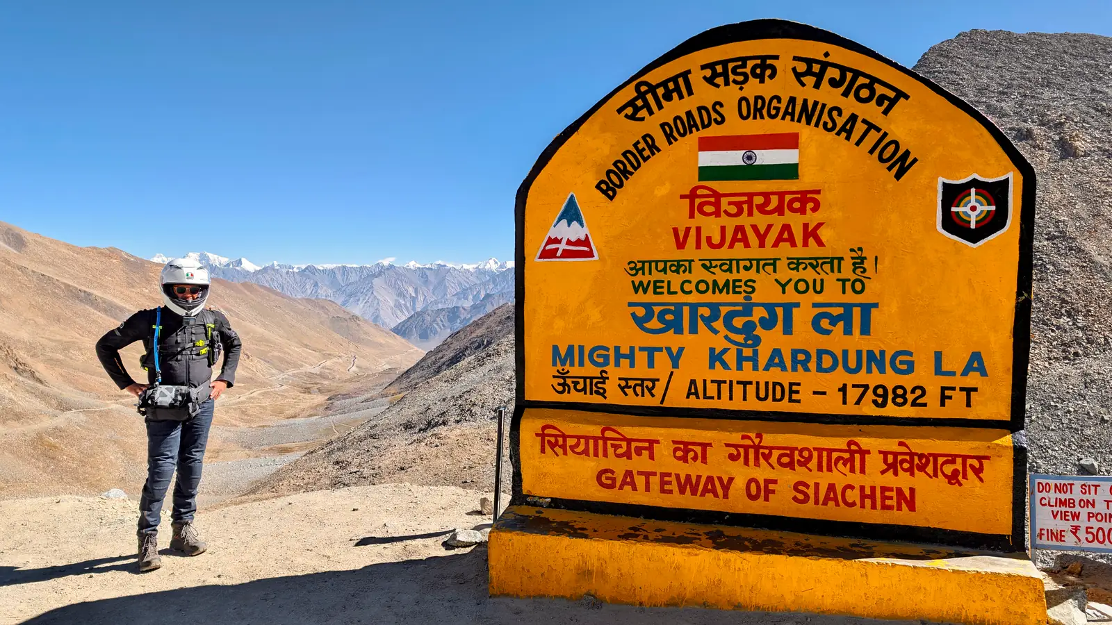 Rider in motorcycle gear standing beside the yellow Khardung La summit sign reading Mighty Khardung La, altitude 17,982 feet, Gateway of Siachen