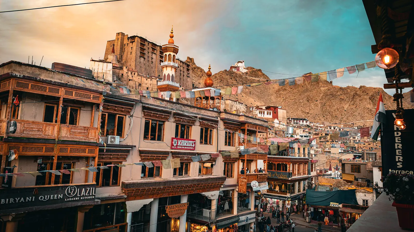 Leh Main Bazaar at dusk, with prayer flags across the street and Leh Palace on the ridge behind