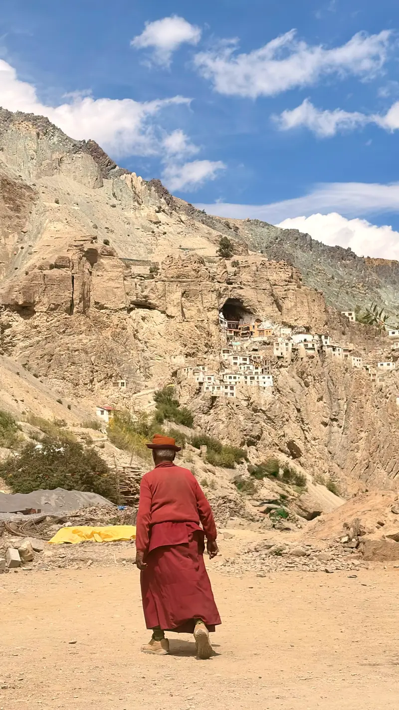 A monk in deep red robes walks the dusty trail toward the monastery village